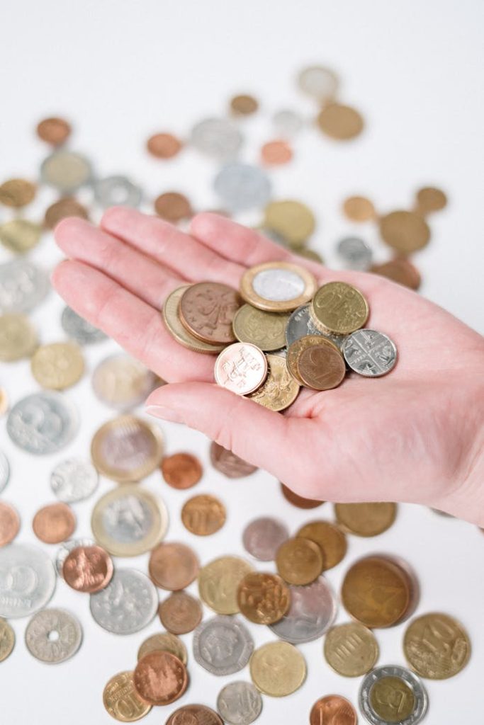 A hand holding a variety of coins with more scattered on a white surface, symbolizing currency and finance.